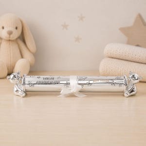 May include: A silver-colored cylindrical birth certificate holder featuring teddy bear figures at each end. The surface is engraved with the text "BIRTH CERTIFICATE" and "Little Joseph" on a banner, accented with star and small icon motifs.