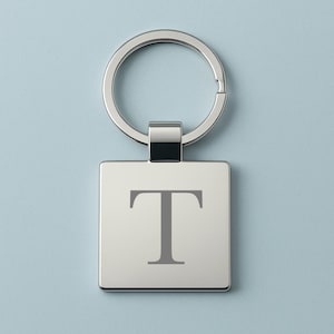 Puede incluir: Llavero de metal plateado con un colgante cuadrado. El colgante presenta la letra "T" gris en una fuente serif. El llavero tiene un anillo partido para sujetar llaves y un conector rectangular.
