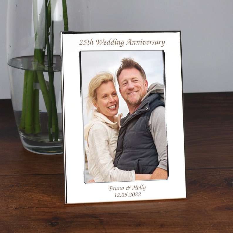 May include: Silver photo frame with a white mat and a picture of a couple. The frame is engraved with "25th Wedding Anniversary", "Bruno & Holly", and "12.05.2022".
