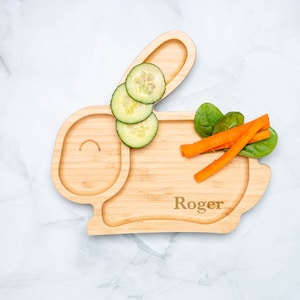 May include: A wooden bunny-shaped divided plate with a name engraved on the bottom. The plate has a section for a main meal and a smaller section for snacks. The plate is filled with cucumber slices, carrot sticks, and spinach leaves.