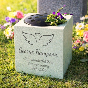 Könnte beinhalten: Ein hellgrüner Gedenkwürfel aus Stein mit einem schwarzen, runden Blumensteck-Einsatz. Die Vorderseite zeigt ein eingraviertes Engelsflügel-Symbol, den Namen George Hampson und den Text Our wonderful Son, Forever young, 1996-2026, mit violetten Blumen.