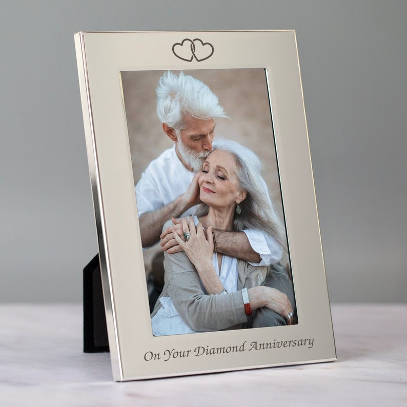 Op de afbeelding: Zilveren fotolijst met twee in elkaar verweven harten gegraveerd aan de bovenkant. De lijst bevat een foto van een ouder koppel dat elkaar omarmt. De onderkant van de lijst is gegraveerd met de tekst "On Your Diamond Anniversary".