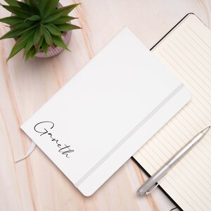 Op de afbeelding: Een wit notitieboek met een zwarte elastische sluiting en de naam "Gwyneth" in cursief op de voorkant. Het notitieboek ligt op een houten oppervlak met een pen en een potplant.
