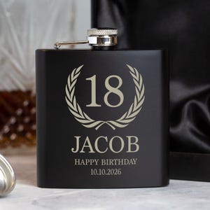 May include: A black hip flask with a silver cap. The front is engraved with the number 18 in a laurel wreath, the name JACOB, and the text "HAPPY BIRTHDAY 10.10.2026". A personalized gift for a special occasion.