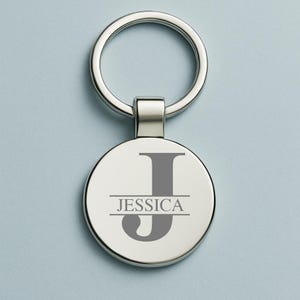 Könnte beinhalten: Ein silberfarbener Schlüsselanhänger mit einem runden Anhänger. Der Anhänger zeigt den Buchstaben "J" in Grau, darunter der Name "JESSICA". Der Schlüsselanhänger hat einen Ring zum Befestigen von Schlüsseln.