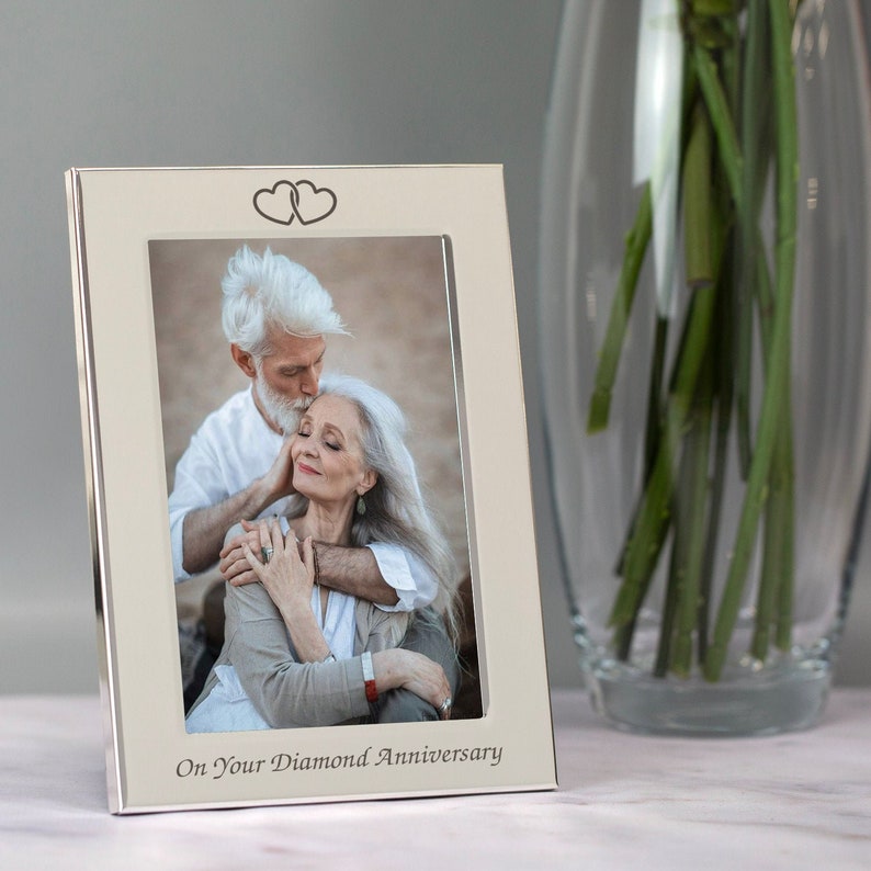 Op de afbeelding: Zilveren fotolijst met een witte passe-partout en twee harten bovenaan. De lijst bevat een foto van een ouder koppel dat elkaar omhelst. De tekst "On Your Diamond Anniversary" is gegraveerd aan de onderkant van de lijst.