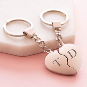 Puede incluir: Un llavero de plata con dos dijes de corazón entrelazados. Los dijes están grabados con las letras "T" y "D".