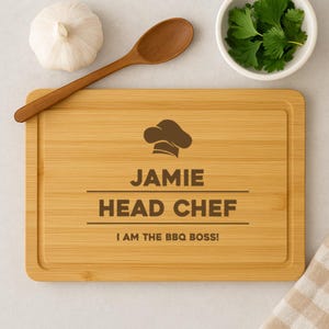 Può includere: Un tagliere in bambù personalizzato con il testo "Jamie Head Chef I am the BBQ Boss!" e un'icona di cappello da cuoco.