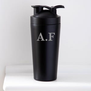 May include: A black shaker bottle with a flip-top lid and a handle. The bottle has the initials "A.F" printed in a light grey colour on the front. The bottle is cylindrical and appears to be made of metal.