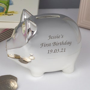 Może przedstawiać: Srebrna skarbonka ze szczeliną na górze. Na skarbonce wygrawerowano tekst "Jessie's First Birthday 19.03.21".