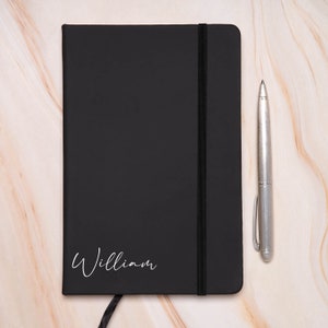 Op de afbeelding: Een zwart lederen notitieboek met een zwarte elastische sluiting en de naam "William" in wit op de voorkant.