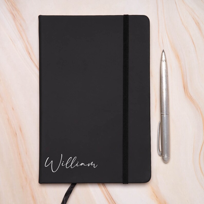 Op de afbeelding: Een zwart lederen notitieboek met een zwarte elastische sluiting en de naam "William" in wit op de voorkant.