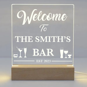 May include: A lighted sign with the words "Welcome To THE SMITH'S BAR" in white script. The sign includes cocktail glasses and the year "EST 2023". The base is made of light-colored wood.