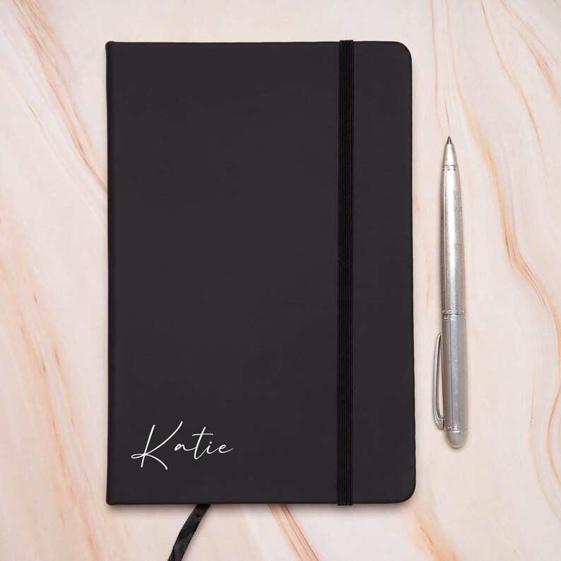 Op de afbeelding: Een zwart notitieboek met een witte elastische sluiting en de naam "Katie" in wit op de voorkant. Een zilveren pen ligt naast het notitieboek op een marmeren oppervlak.