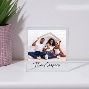 May include: A clear glass photo block with a family photo inside. The photo shows a family of four, two parents and two children, sitting in front of a white brick wall. The parents are holding a white paper house over their heads. The text "The Coopers" is printed on the bottom of the block.