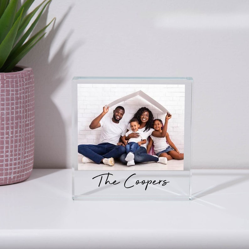 May include: A clear glass photo block with a family photo inside. The photo shows a family of four, two parents and two children, sitting in front of a white brick wall. The parents are holding a white paper house over their heads. The text "The Coopers" is printed on the bottom of the block.