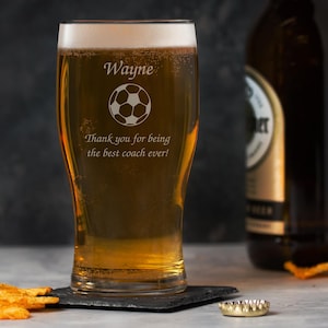 May include: A pint glass filled with golden beer, engraved with the name "Wayne", a soccer ball, and the message "Thank you for being the best coach ever!". A beer bottle, bottle cap, and snacks are also visible.