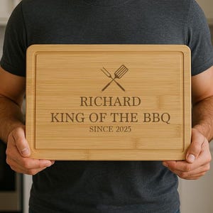 Op de afbeelding: Een rechthoekige bamboe snijplank met een sapgroef. De plank heeft de tekst "RICHARD KING OF THE BBQ SINCE 2025" met een gekruist vork- en spatelontwerp. De plank is lichtbruin.