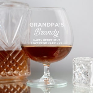 May include: A clear glass brandy snifter with the text "Grandpa's Brandy" and "Happy Retirement Love from Katie xxx" etched on the side. The glass is filled with amber-colored brandy.