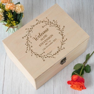Op de afbeelding: Een houten bewaardoos van licht hout met een gegraveerd ontwerp van een bloemkrans. De tekst luidt "Welkom Alfie Stevenson 6lb 7oz 12 november 2023".