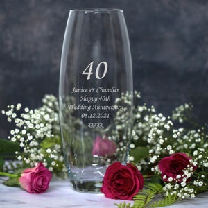 Può includere: Un vaso di vetro trasparente con il numero 40 inciso sul davanti. Il vaso è decorato con il testo "Janice & Chandler Happy 40th Wedding Anniversary 08.12.2021". Il vaso è circondato da fiori bianchi e rosa.