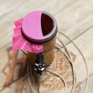 May include: A wooden container with a pink rubber cover and a black band. A decorative cord with a black and white bead hangs from the container. The container is sitting on a light brown surface.