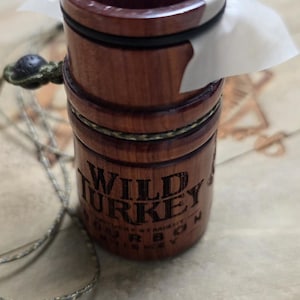 May include: A wooden Wild Turkey bourbon bottle stopper with a paper seal. The stopper is cylindrical, with a dark brown finish and black accents. The text "WILD TURKEY" is visible on the side. A green and gray cord is attached.