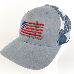 May include: A gray and blue baseball cap with a mesh back. The front of the cap features an embroidered American flag with a cross and the words "In God We Trust".