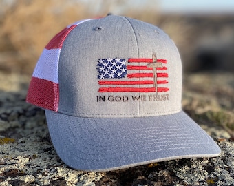 May include: A gray and red baseball cap with a white mesh back. The front of the cap features an embroidered American flag with a cross and the words "In God We Trust".