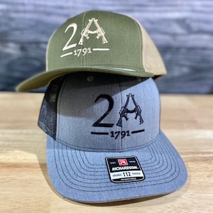 May include: Two trucker hats stacked on top of each other. The top hat is olive green with a tan mesh back and features a white embroidered design of two crossed rifles with the text "2A -1791-". The bottom hat is gray with a black mesh back and features a black embroidered design of two crossed rifles with the text "2A -1791-". The bottom hat also has a black label with the text "RICHARDSON. ORIGINAL 112 TRUCKER ADJUSTABLE".