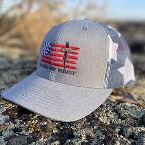 May include: A gray and white trucker hat with a red, white, and blue American flag design and the words "In God We Trust" embroidered on the front.