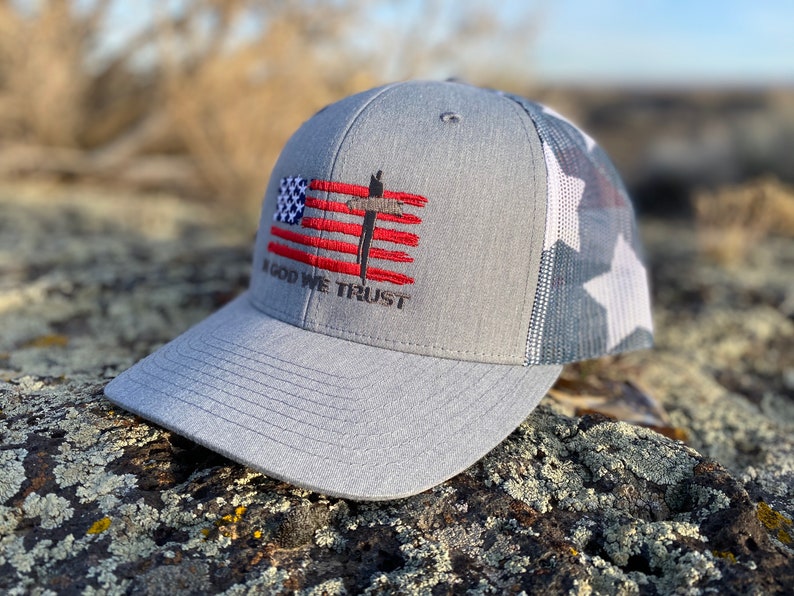 May include: A gray and white trucker hat with a red, white, and blue American flag design and the words "In God We Trust" embroidered on the front.