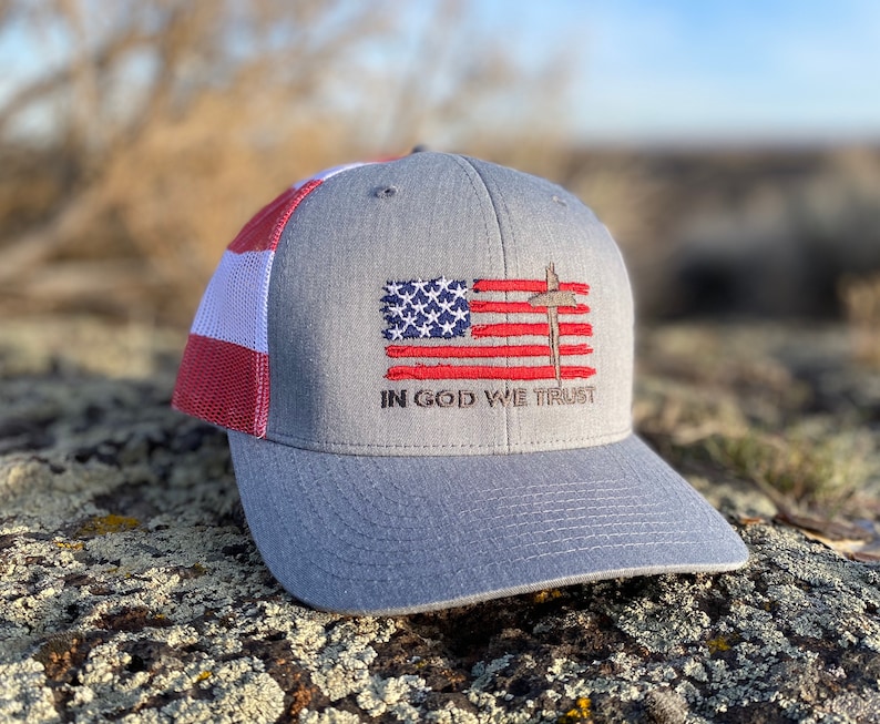 May include: A gray and red baseball cap with a white mesh back. The front of the cap features an embroidered American flag with a cross and the words "In God We Trust".