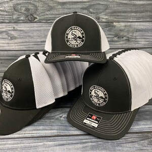 Może przedstawiać: Stos czarno-białych czapek bejsbolowych z białym siateczkowym tyłem. Czapki mają białe haftowane logo z napisem "Wai-Kamala White Sox".