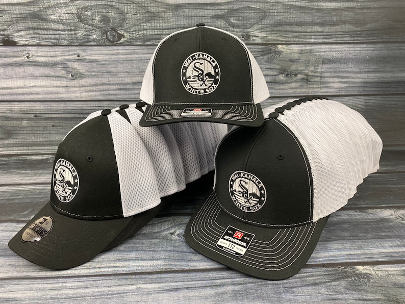 Może przedstawiać: Stos czarno-białych czapek bejsbolowych z białym siateczkowym tyłem. Czapki mają białe haftowane logo z napisem "Wai-Kamala White Sox".