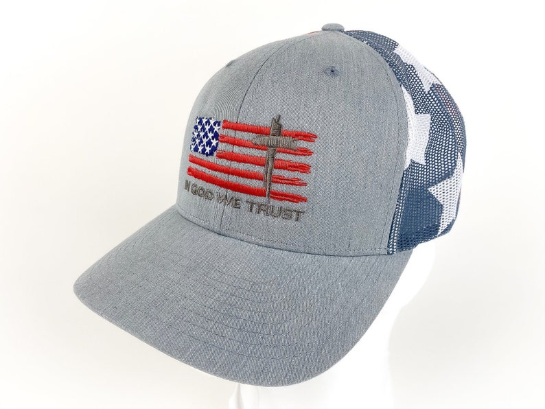May include: A gray and blue baseball cap with a mesh back. The front of the cap features an embroidered American flag with a cross and the words "In God We Trust".