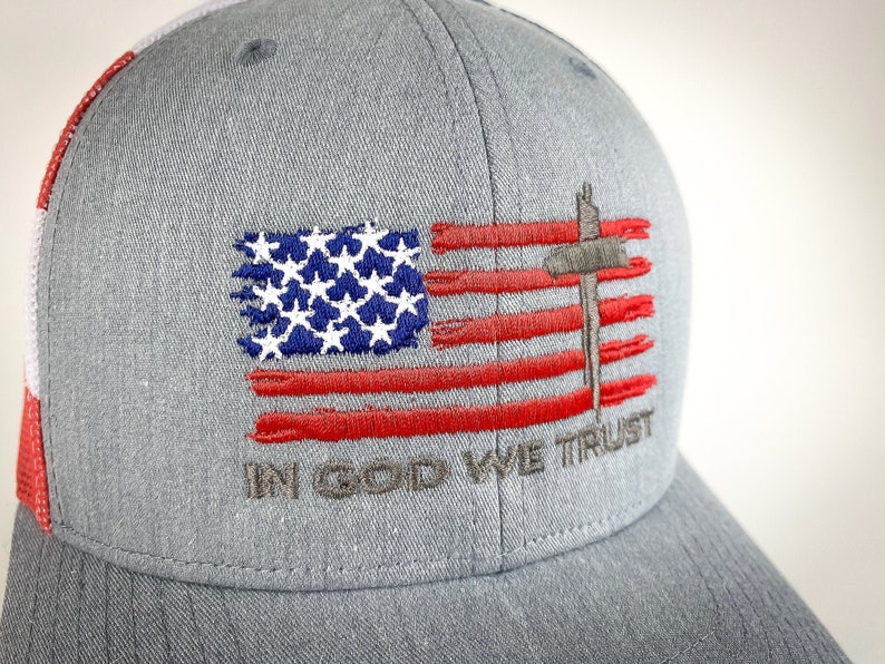 May include: A gray and red baseball cap with an embroidered American flag and the words "In God We Trust".