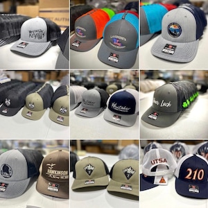 May include: A collection of various embroidered baseball caps in different colors and designs. The caps feature a variety of logos and text, including "Street Royalty", "LaVergne's", "Whitaker", "Cheer Luck", "Hawkinson", "UTSA", and "210".