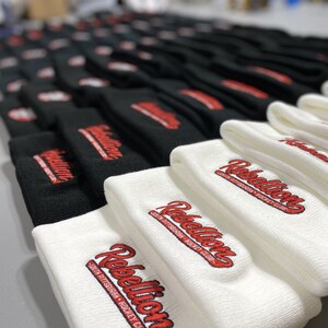 May include: A stack of white and black knit beanies with the embroidered text "Rebellion South Pittsburgh Hockey Club" in red.