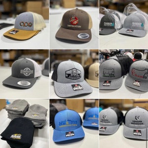 May include: A collection of embroidered hats with various logos and designs. The hats are a mix of gray, brown, blue, and black. Some of the logos include "Dirtbusters", "Marker Surf Shop", "Cornerstone", "Layman Cattle", "C Farms", "Soul & Terre Supernatural Scents", and "Upstate Woodwork".