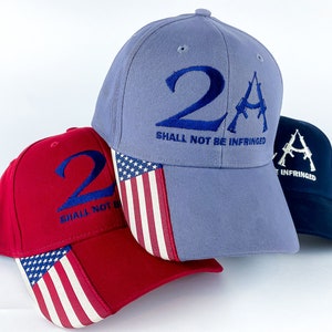 May include: Three baseball caps with an American flag design. The caps are blue, red, and navy blue. The front of each cap has the number "2" and the letter "A" with a stylized image of a gun. The text "Shall not be infringed" is printed below the number and letter on the blue and red caps. The navy blue cap has the text "Shall not be infringed" printed below the letter "A".
