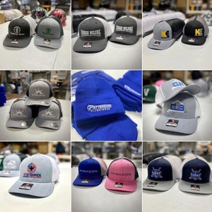 May include: A collection of various embroidered hats with different logos and designs. The hats are in different colors, including gray, black, blue, pink, and white. Some of the logos include "Patterson Plumbing", "13 Stripes", "Paragon", and "Count".