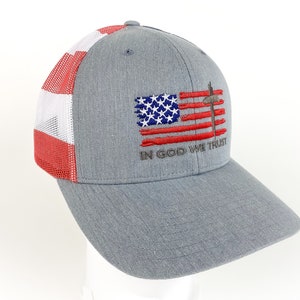May include: A gray and red trucker hat with a mesh back. The front of the hat features an embroidered American flag with a cross and the words "In God We Trust".
