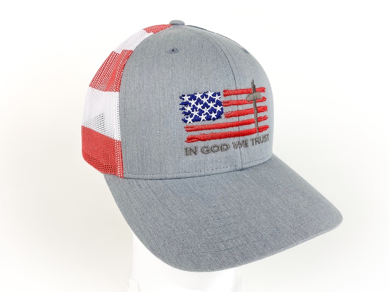 May include: A gray and red trucker hat with a mesh back. The front of the hat features an embroidered American flag with a cross and the words "In God We Trust".