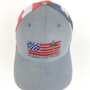 May include: A gray baseball cap with a red, white, and blue mesh back. The front of the cap features an embroidered American flag with a cross and the text "In God We Trust".