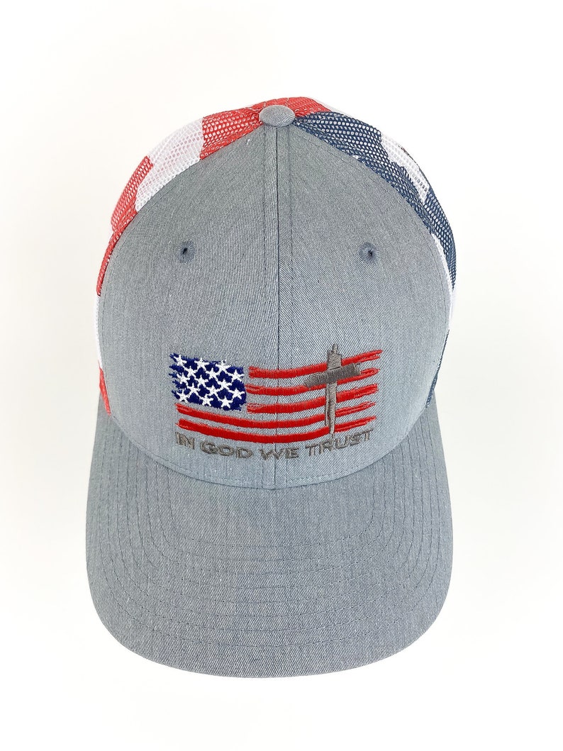 May include: A gray baseball cap with a red, white, and blue mesh back. The front of the cap features an embroidered American flag with a cross and the text "In God We Trust".