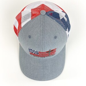 May include: A gray and blue mesh trucker hat with a red, white, and blue American flag design on the front. The flag has a cross on it and the words "God First" are embroidered below it.