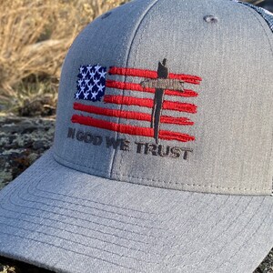 May include: A gray and white baseball cap with a red, white, and blue American flag design. The flag has a brown cross in the center. The text "In God We Trust" is embroidered below the flag.