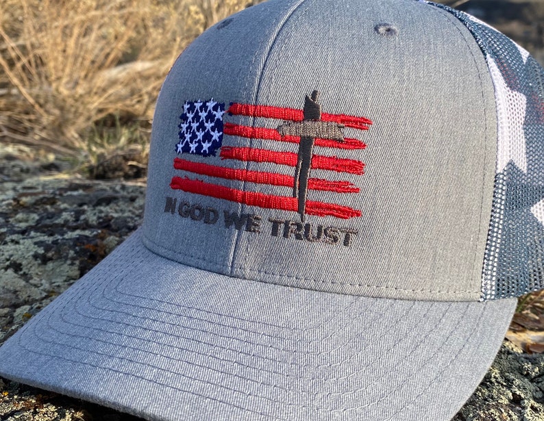 May include: A gray and white baseball cap with a red, white, and blue American flag design. The flag has a brown cross in the center. The text "In God We Trust" is embroidered below the flag.