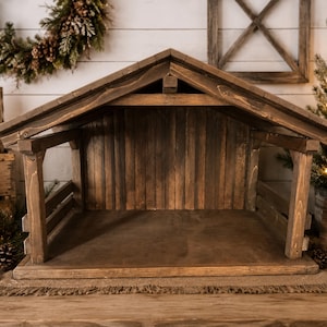 May include: A dark brown wooden nativity stable featuring a gabled roof, vertical plank back wall, and open sides with horizontal slats. The structure is constructed from stained wood with visible grain, designed as a display for figurines.